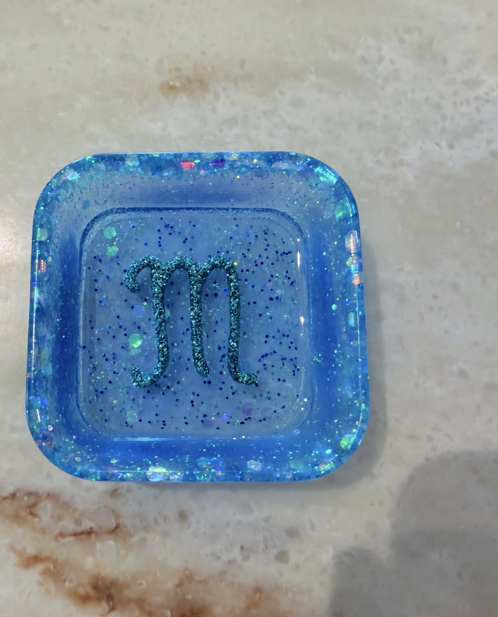 Square rounded translucent blue resin dish filled with iridescent glitter and a teal glitter script lowercase m in the center